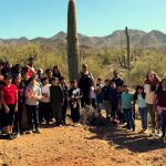 Teacher and large group of students stand in front of saguaros and mountain landscape at Saguaro National Park