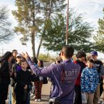 A group of students circle around and listen to a community partner while standing in a park with large trees in the background.
