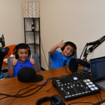 Two students give a thumbs up with headphones on while sitting at podcast recording studio
